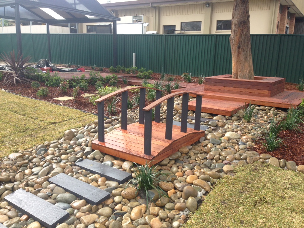 Small timber footbridge over a dry creek bed in a finished backyard landscape.